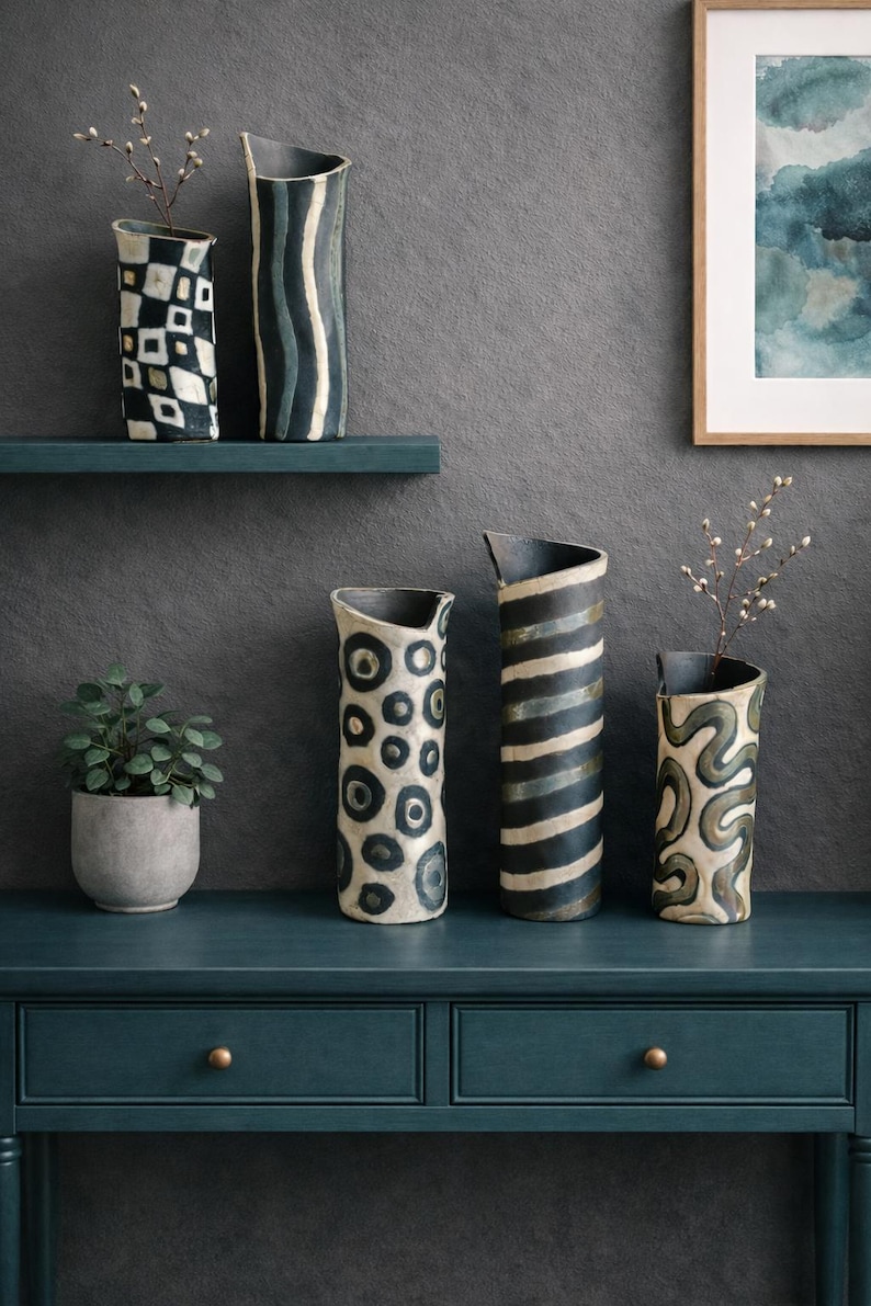 May include: A collection of ceramic vases with unique patterns, including geometric shapes, stripes, and organic designs. The vases are in shades of white, black, and gray, displayed on a teal console table and shelf. A small potted plant and framed artwork complete the decor.