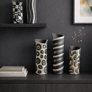 May include: A collection of ceramic vases with unique patterns. The vases feature designs such as black and white geometric shapes, stripes, and organic swirls. Some vases hold small branches, adding a natural element to the display.