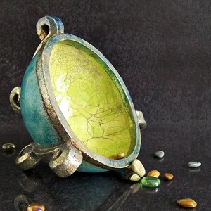 May include: A handcrafted, teal-colored ceramic bowl with a textured, crackled yellow interior. The bowl has a silver rim and base, with small decorative handles. Small, colorful stones are scattered around the bowl.