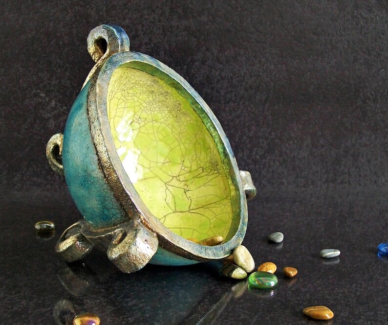 May include: A handcrafted, teal-colored ceramic bowl with a textured, crackled yellow interior. The bowl has a silver rim and base, with small decorative handles. Small, colorful stones are scattered around the bowl.