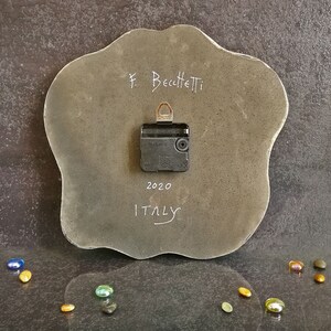 Pu&ograve; includere: Il retro di un orologio da parete grigio di forma irregolare con un meccanismo nero a batteria. La superficie presenta la scritta F. Becchetti, 2020 e ITALY in bianco. Un gancio metallico &egrave; fissato nella parte superiore del meccanismo.