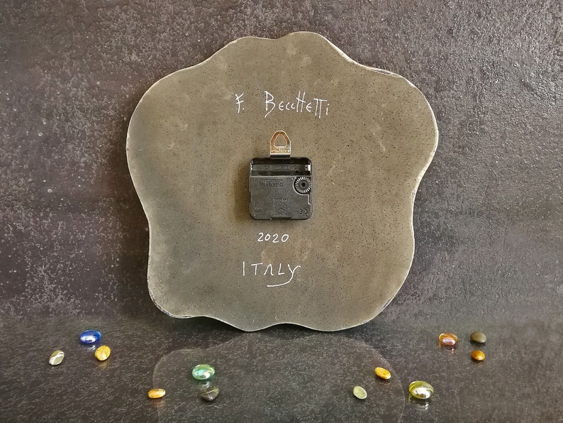 Pu&ograve; includere: Il retro di un orologio da parete grigio di forma irregolare con un meccanismo nero a batteria. La superficie presenta la scritta F. Becchetti, 2020 e ITALY in bianco. Un gancio metallico &egrave; fissato nella parte superiore del meccanismo.