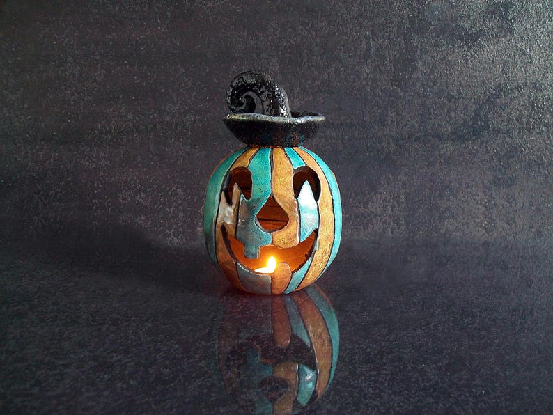 May include: A ceramic jack-o'-lantern candle holder featuring vertical stripes in shades of teal and tan. The pumpkin has a carved face with triangular eyes and a jagged mouth, topped with a black, curled, hat-like stem. A warm light glows from within.