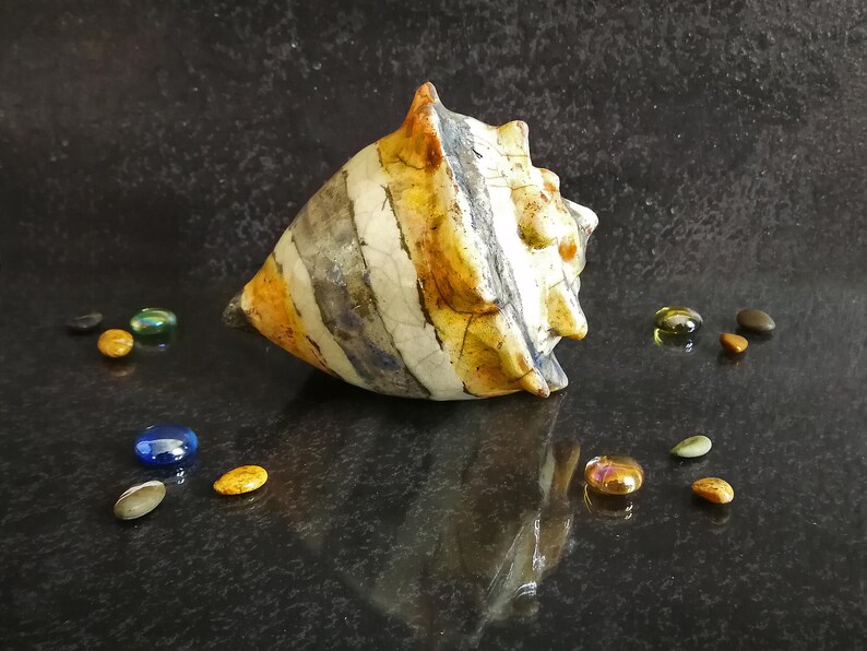 Puede incluir: Una concha de cer&aacute;mica con un patr&oacute;n a rayas en tonos blancos, naranjas y grises. La concha est&aacute; rodeada de peque&ntilde;os guijarros de vidrio de colores sobre una superficie oscura y reflectante. Objeto decorativo.
