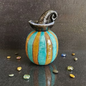 May include: A ceramic pumpkin-shaped jar with a glossy finish, featuring alternating vertical stripes of turquoise and mustard yellow. The lid is topped with a dark, curved, textured stem handle, creating a distinct contrast with the body.