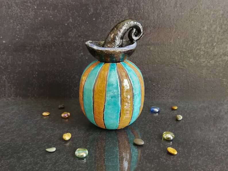 May include: A ceramic pumpkin-shaped jar with a glossy finish, featuring alternating vertical stripes of turquoise and mustard yellow. The lid is topped with a dark, curved, textured stem handle, creating a distinct contrast with the body.