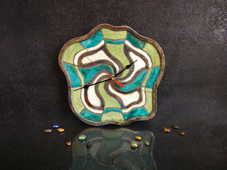 May include: A ceramic clock with an abstract design in green, blue and white. The clock has a black hour hand and a red minute hand.