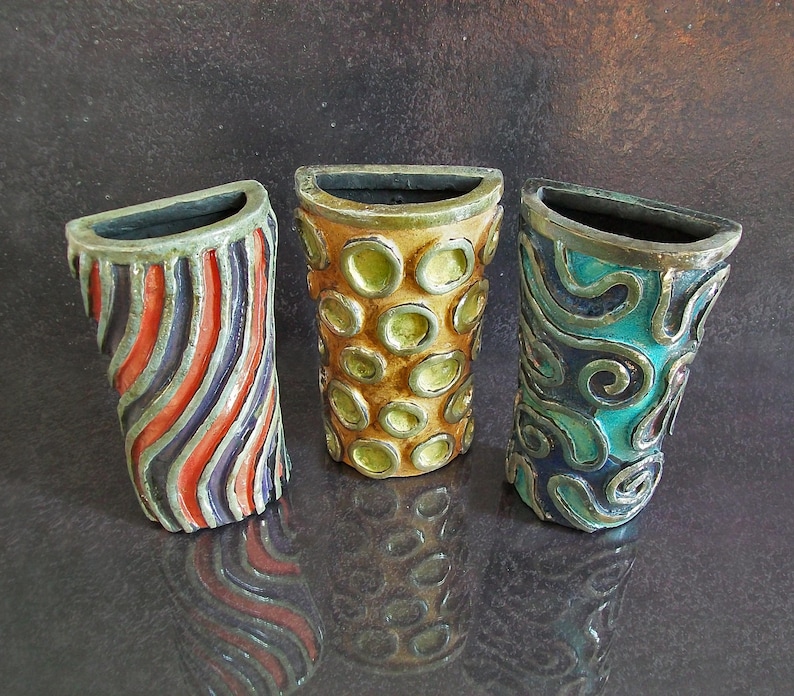 May include: Three decorative ceramic vases with a unique design. One vase features red, blue, and gray diagonal stripes. Another has a pattern of yellow circles on a brown background. The third vase has a teal and dark blue swirl design.