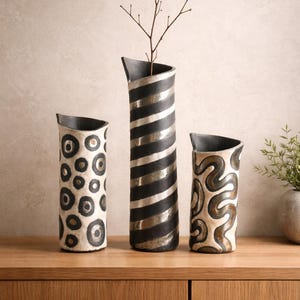 May include: Three decorative ceramic vases with unique designs. The tallest vase has black and silver stripes, the middle vase has a pattern of black circles, and the smallest vase has a brown and cream swirl design. A twig is in the tallest vase.