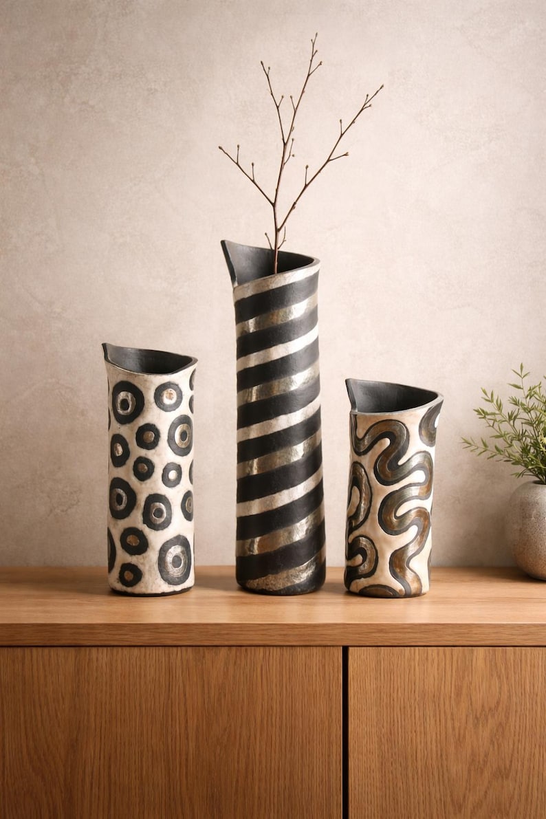 May include: Three decorative ceramic vases with unique designs. The tallest vase has black and silver stripes, the middle vase has a pattern of black circles, and the smallest vase has a brown and cream swirl design. A twig is in the tallest vase.