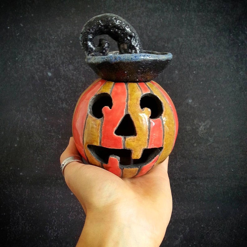 May include: A ceramic jack-o'-lantern with alternating vertical orange and yellow stripes, featuring a carved face with two round eyes, a triangular nose, and a wide mouth. A black, textured, curled stem sits on top of the round pumpkin body.