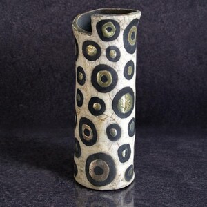 May include: A cylindrical ceramic vase with a cream-colored base and a pattern of black and gold circles. The vase has a slightly curved top and a dark interior. The design is repeated around the vase, creating a visually interesting pattern.