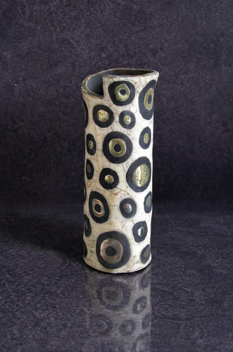 May include: A cylindrical ceramic vase with a cream-colored base and a pattern of black and gold circles. The vase has a slightly curved top and a dark interior. The design is repeated around the vase, creating a visually interesting pattern.