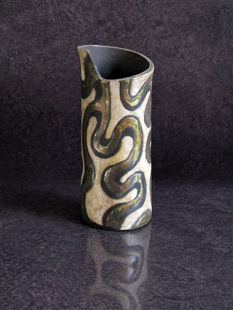 May include: A ceramic pitcher with a cream-colored base and a black and gold swirling pattern. The top of the pitcher is black, and the design is reminiscent of abstract art. The pitcher is displayed on a dark surface.