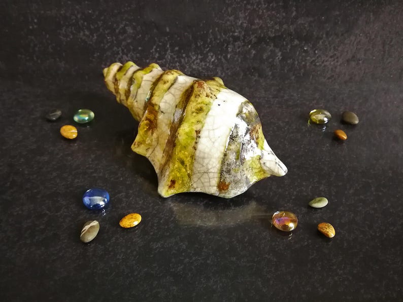 Puede incluir: Una escultura de concha de cer&aacute;mica con una combinaci&oacute;n de colores crema, verde y marr&oacute;n. La concha est&aacute; rodeada de peque&ntilde;os guijarros de vidrio de colores. La escultura tiene una superficie texturizada y es un objeto decorativo.