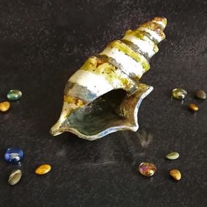 Puede incluir: Una escultura de concha de cer&aacute;mica con un dise&ntilde;o en espiral, con rayas blancas, verdes y amarillas. La concha est&aacute; rodeada de peque&ntilde;as piedras decorativas de colores. La escultura est&aacute; sobre una superficie oscura y texturizada.