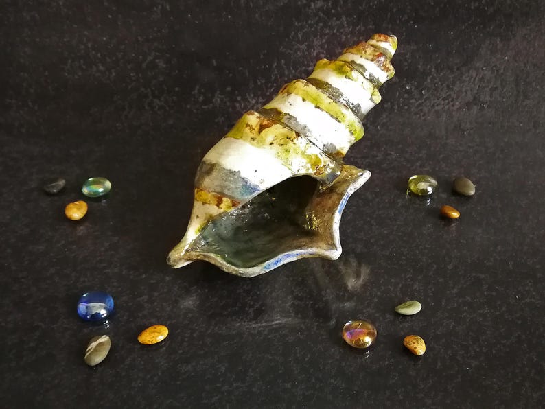 Puede incluir: Una escultura de concha de cer&aacute;mica con un dise&ntilde;o en espiral, con rayas blancas, verdes y amarillas. La concha est&aacute; rodeada de peque&ntilde;as piedras decorativas de colores. La escultura est&aacute; sobre una superficie oscura y texturizada.
