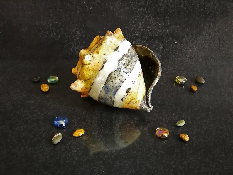 Puede incluir: Una concha de cer&aacute;mica con rayas amarillas, blancas y grises, sobre una superficie oscura. Peque&ntilde;as piedras de colores est&aacute;n dispersas alrededor de la concha. La abertura de la concha revela un interior oscuro.