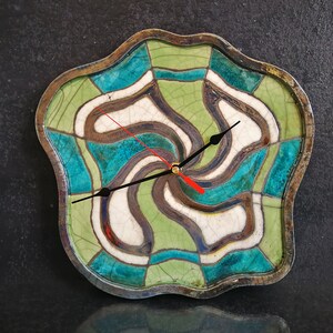 Pu&ograve; includere: Un orologio da parete con una cornice in ceramica ondulata e irregolare con finitura smaltata a crepa. Il quadrante presenta un motivo geometrico vorticoso nei toni del verde salvia, turchese e bianco, accentuato da linee marrone scuro. Ha lancette nere e una lancetta dei secondi rossa.