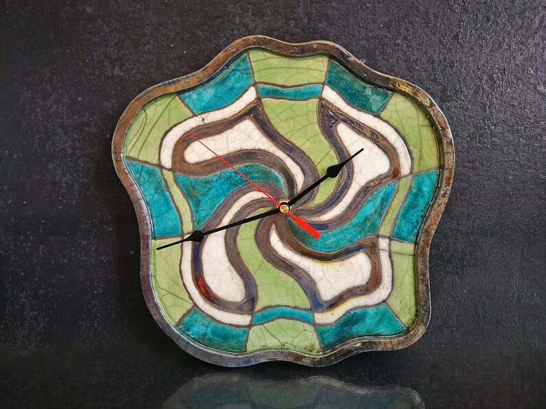 Pu&ograve; includere: Un orologio da parete con una cornice in ceramica ondulata e irregolare con finitura smaltata a crepa. Il quadrante presenta un motivo geometrico vorticoso nei toni del verde salvia, turchese e bianco, accentuato da linee marrone scuro. Ha lancette nere e una lancetta dei secondi rossa.