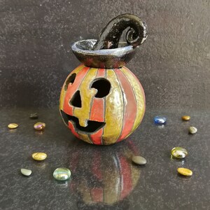 May include: A ceramic jack-o'-lantern candle holder featuring vertical red and yellow stripes, a glossy finish, and a black, curled stem on top. The pumpkin has cutouts for eyes, a nose, and a mouth, with a reflective surface and scattered glass beads.
