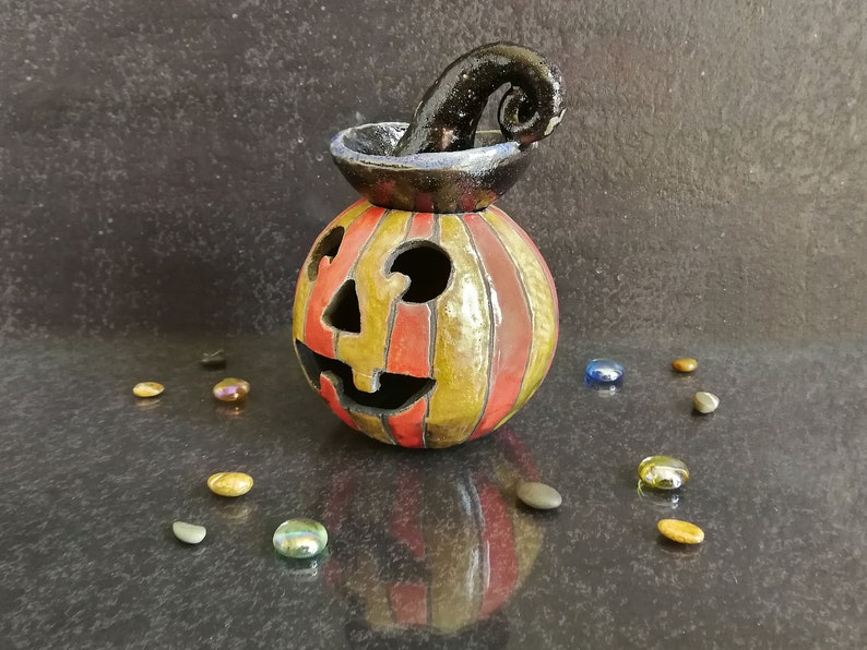 May include: A ceramic jack-o'-lantern candle holder featuring vertical red and yellow stripes, a glossy finish, and a black, curled stem on top. The pumpkin has cutouts for eyes, a nose, and a mouth, with a reflective surface and scattered glass beads.