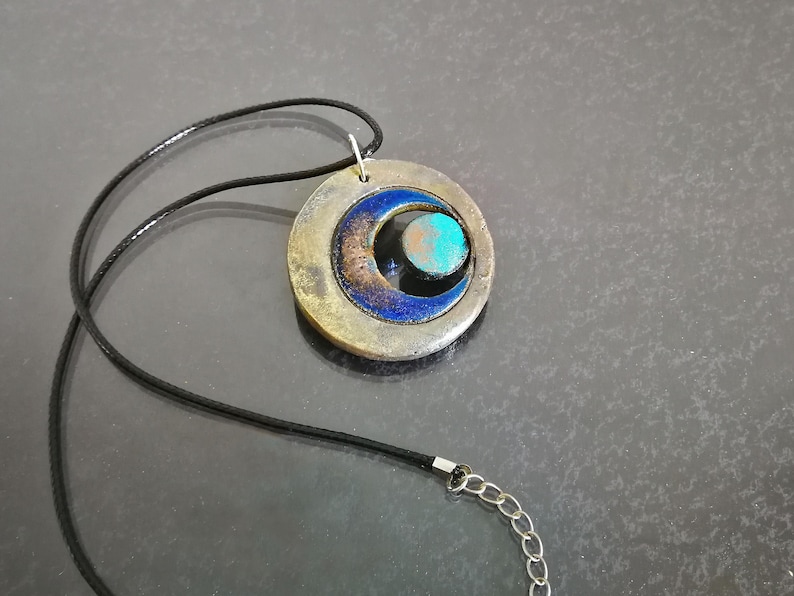 May include: A pendant necklace with a circular, mixed-metal pendant. The pendant has a crescent shape and a turquoise-coloured stone. It hangs from a black cord with a silver-coloured chain clasp. The design includes blue and bronze tones.