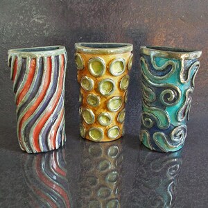 May include: Three decorative ceramic vases with unique designs. One vase features red, orange, and purple diagonal stripes. Another has a yellow background with green circles. The third vase has a teal background with silver swirls.