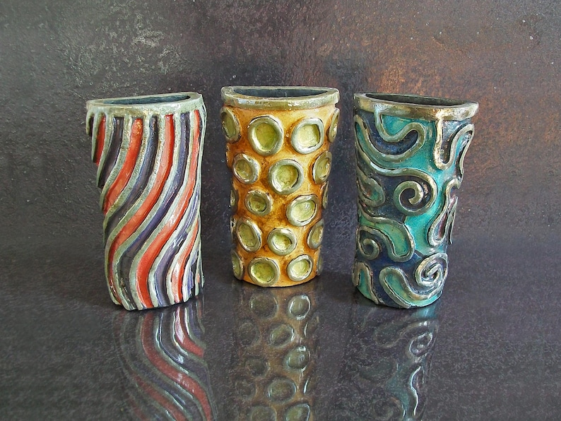 May include: Three decorative ceramic vases with unique designs. One vase features red, orange, and purple diagonal stripes. Another has a yellow background with green circles. The third vase has a teal background with silver swirls.