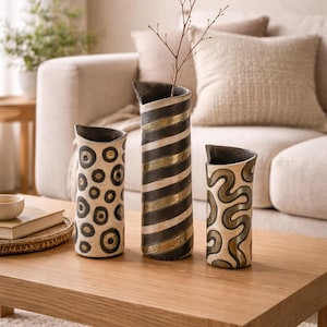 May include: Three decorative ceramic vases with unique patterns. One vase features black and gold stripes, another has a pattern of black circles, and the third has a swirling design. The vases are displayed on a wooden coffee table.