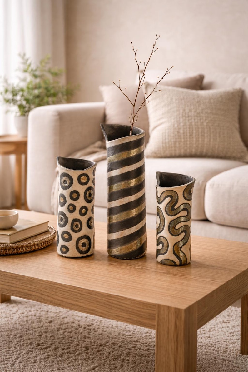May include: Three decorative ceramic vases with unique patterns. One vase features black and gold stripes, another has a pattern of black circles, and the third has a swirling design. The vases are displayed on a wooden coffee table.