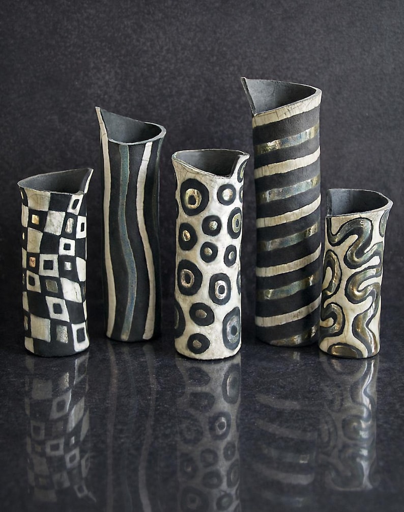 May include: Five handcrafted ceramic vases with unique black and white patterns. Designs include geometric shapes, wavy lines, and circular patterns with metallic accents. Each vase has a distinct shape and a dark gray interior.