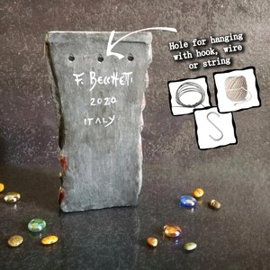May include: A rectangular, gray stone art piece with three holes for hanging. The text "F. Becchetti 2020 ITALY" is inscribed on the back. Decorative glass stones in various colors are scattered around the base.