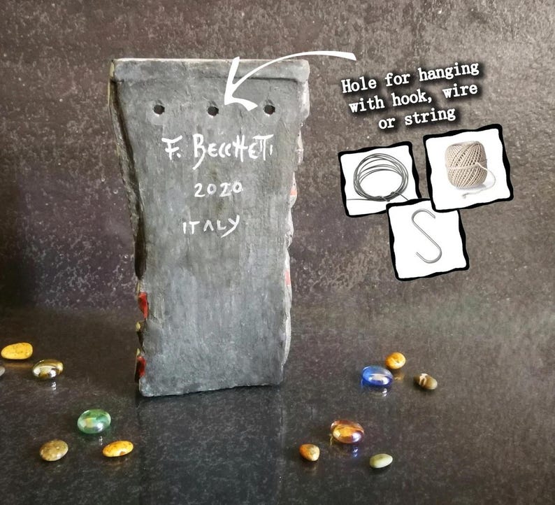 May include: A rectangular, gray stone art piece with three holes for hanging. The text "F. Becchetti 2020 ITALY" is inscribed on the back. Decorative glass stones in various colors are scattered around the base.