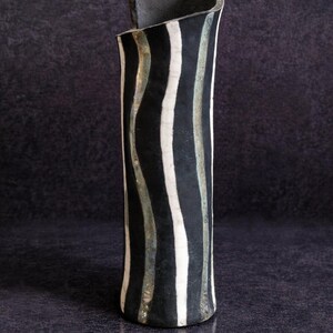 May include: A tall, cylindrical ceramic vase with a black and white striped pattern. The vase has a curved top and a dark, textured finish. The stripes are vertical and wavy, creating a unique design.