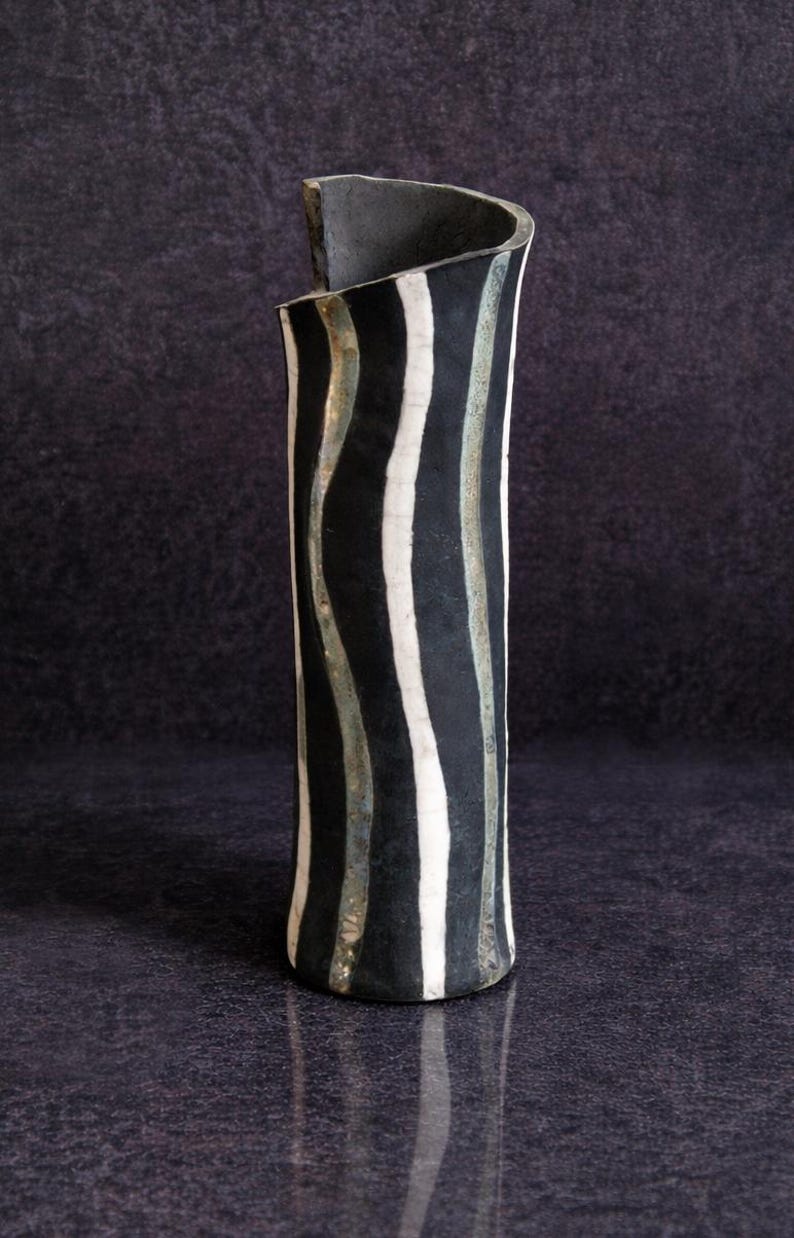 May include: A tall, cylindrical ceramic vase with a black and white striped pattern. The vase has a curved top and a dark, textured finish. The stripes are vertical and wavy, creating a unique design.