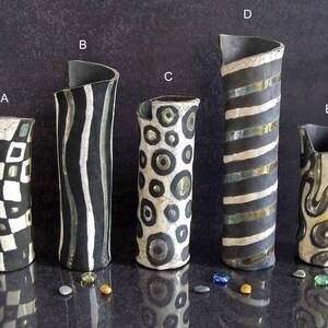 May include: Five handcrafted ceramic vases with unique designs. The vases feature black and white patterns including squares, stripes, and circles. Each vase has a distinctive shape and a dark, textured finish. Decorative pebbles are scattered around the vases.