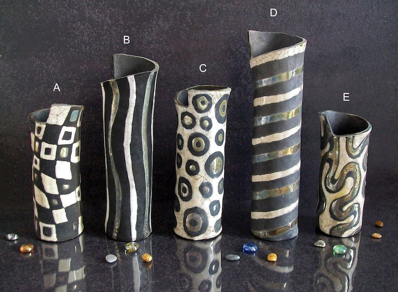 May include: Five handcrafted ceramic vases with unique designs. The vases feature black and white patterns including squares, stripes, and circles. Each vase has a distinctive shape and a dark, textured finish. Decorative pebbles are scattered around the vases.