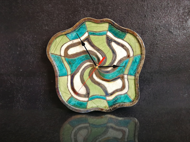 May include: A ceramic clock with a green, blue and white abstract design. The clock has a wavy, irregular shape and black hands.
