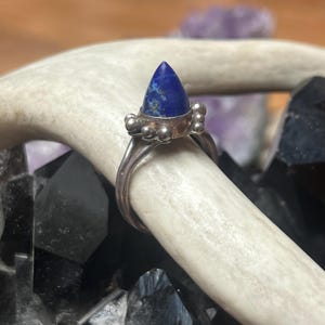 May include: A silver ring with a blue lapis lazuli stone set in a pointed shape. The ring has a textured band and a row of small silver beads around the stone.