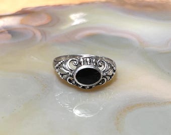 May include: A silver ring with an oval black stone. The ring has an ornate design with scrollwork and decorative elements around the stone. The ring is set against a neutral background.