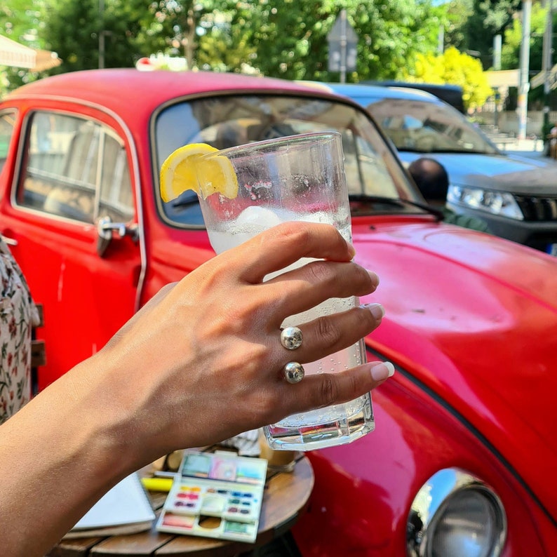 以下が含まれることがあります： レモンのくさび形にカットされたものを添えたレモネードが入ったグラスを手に持っている。グラスは赤いビンテージカーの上に置かれている。