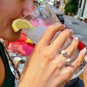 以下が含まれることがあります： レモンのくさび形が置かれた透明な液体が入ったグラスから飲んでいる人。その人は、2つの丸いボールが付いた銀の指輪をしています。