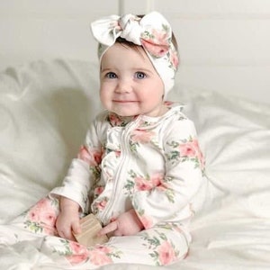 May include: A baby wearing a white floral print onesie with a ruffle trim and a matching floral headband. The onesie has a zipper closure and long sleeves.