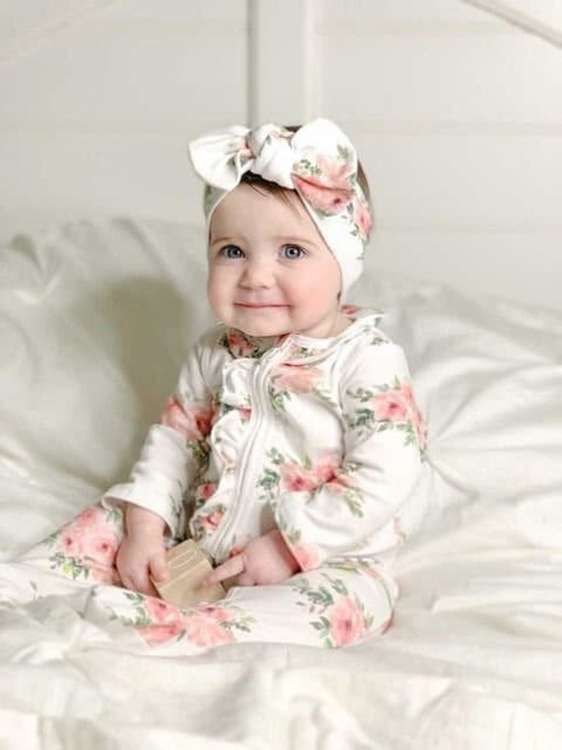 May include: A baby wearing a white floral print onesie with a ruffle trim and a matching floral headband. The onesie has a zipper closure and long sleeves.
