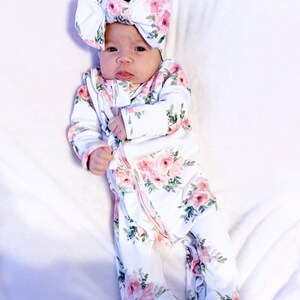 May include: A baby wearing a white floral print sleeper with a pink floral headband. The sleeper has a zipper closure and ruffles at the bottom.