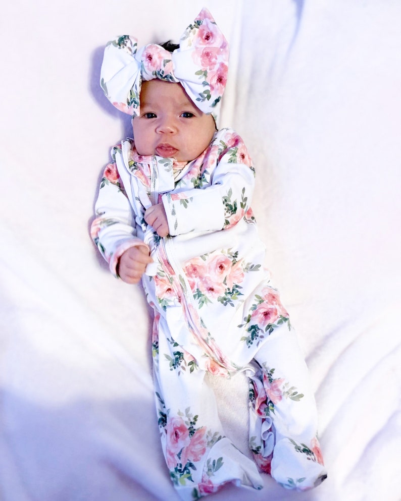 May include: A baby wearing a white floral print sleeper with a pink floral headband. The sleeper has a zipper closure and ruffles at the bottom.