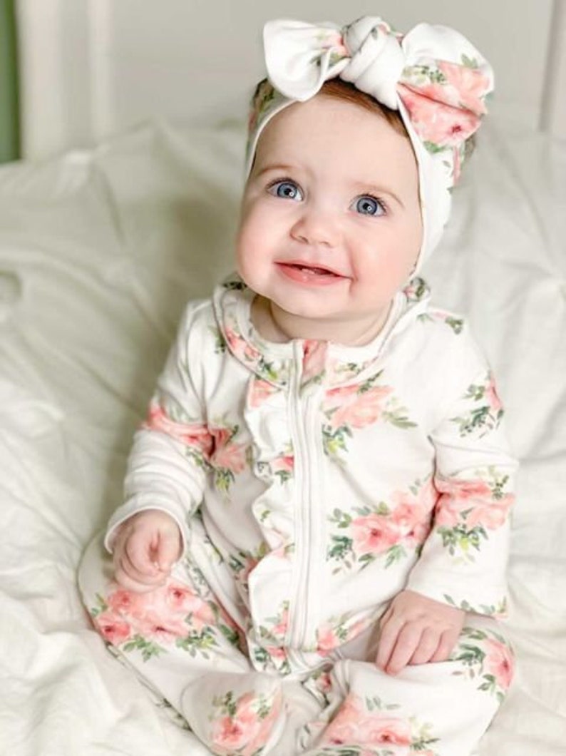 May include: A baby wearing a white floral print long-sleeved onesie with a ruffled neckline and a white headband with a pink floral print.