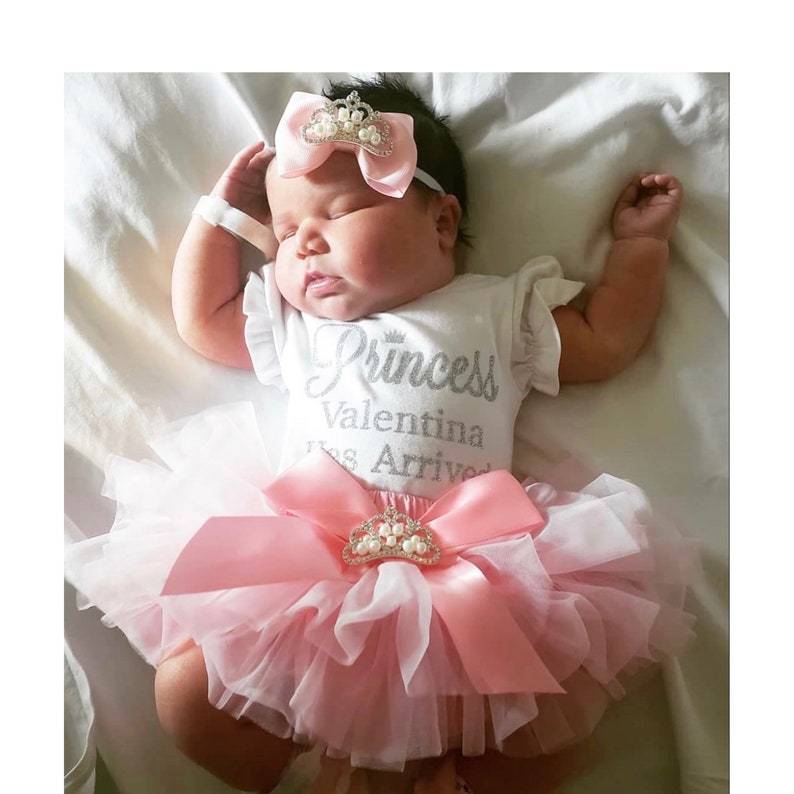 May include: A baby girl wearing a white onesie with the text "Princess Valentina Has Arrived" in silver glitter. She is wearing a pink tulle tutu with a pink satin bow and a silver crown with pearls. The baby is sleeping on a white blanket.