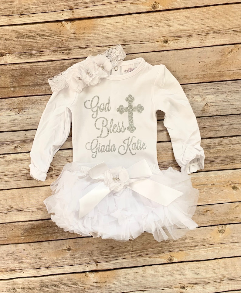 May include: White baby outfit with long sleeves and a tulle skirt. The shirt has a silver glitter design that says "God Bless Giada Katie" with a cross.